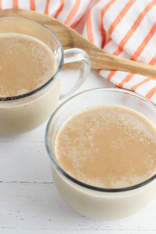 This photo features a two cups of pumpkin spiced latte sitting on a white bead board table, next to it is a orange and white striped tea towel and wooden spoon.