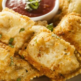 A plate of golden-brown baked ravioli appetizers with a white side dish of pasta sauce for dipping.