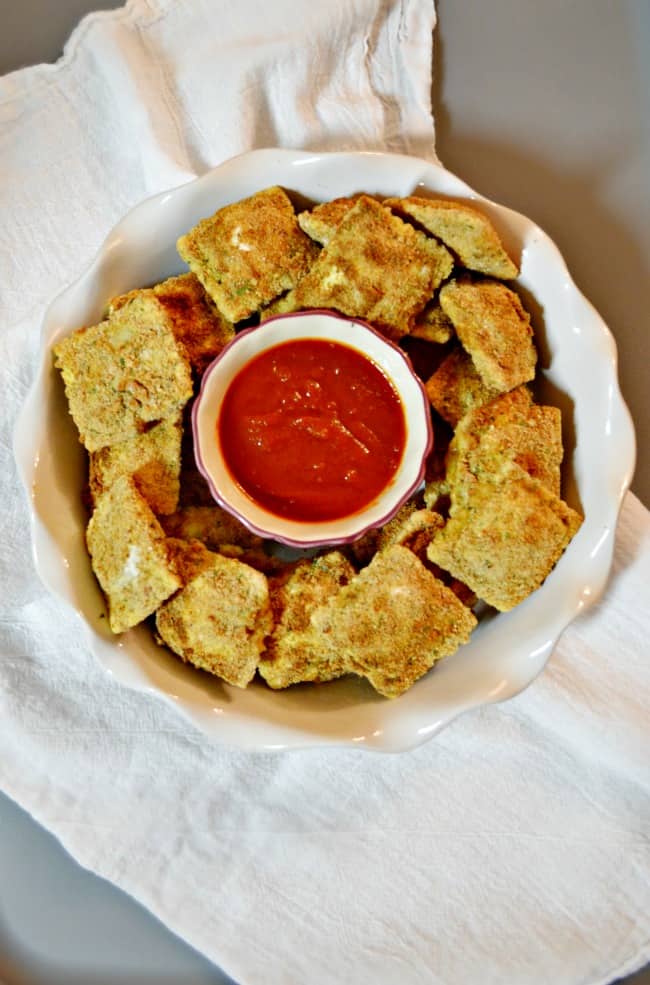 A large white bowl filled with the baked ravioli appetizers with a small bowl of warmed sauce in the center.