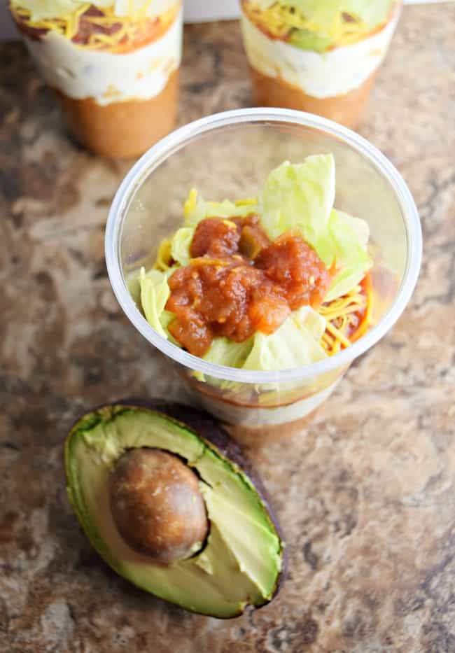 Photo of the 7 layer dip in a plastic cup with a sliced in half avocado including the pit.