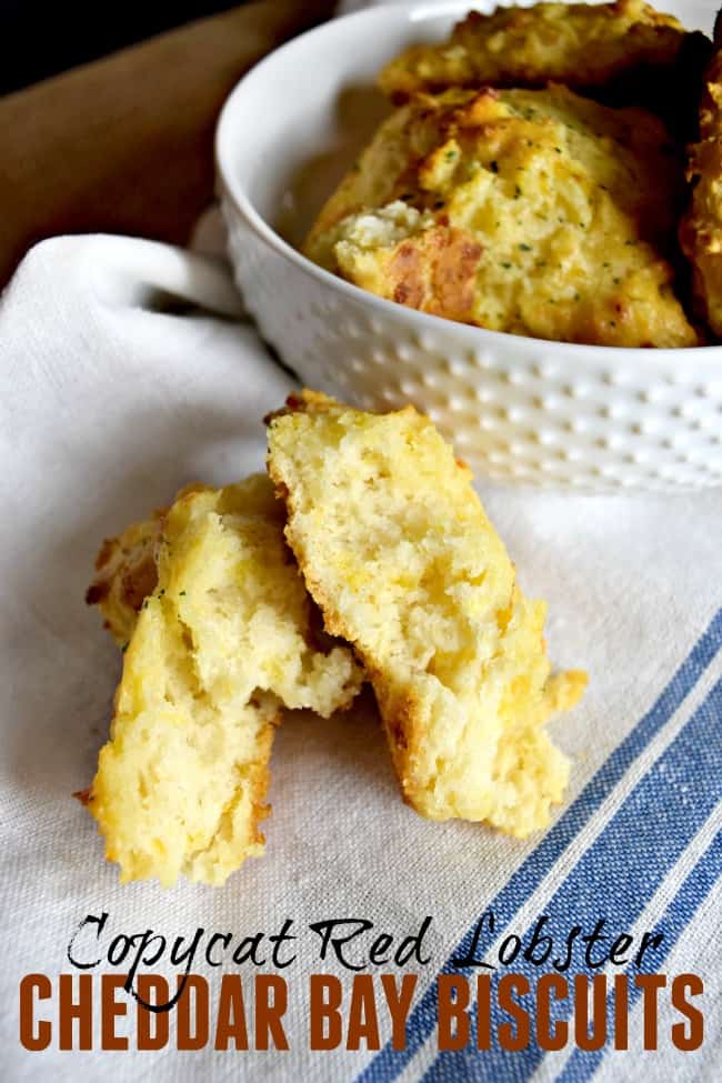 A pile of Red Lobster Cheddar Bay Biscuits in a white bowl and one sitting on a tea towel that is broken into.