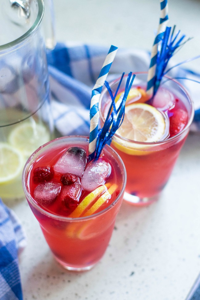 This photo features a summer berry lemonade recipe. There is 2 glasses filled with the beverage with ice cubes, and extra fruit in the glass. There is a blue and white straw in the glass and they are sitting next to a pitcher of the drink.