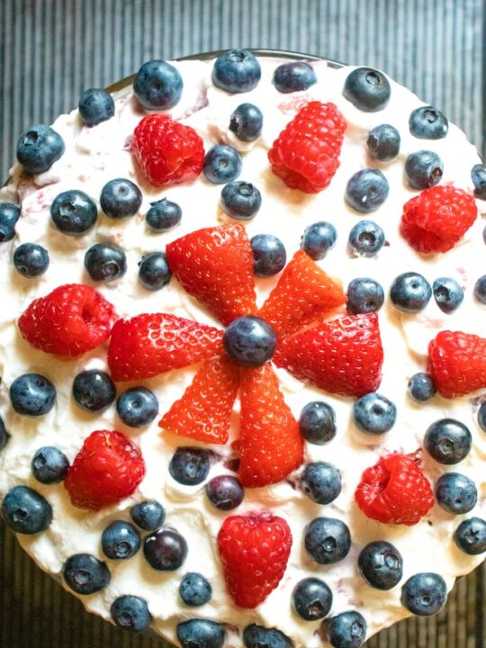 This photo features a above angle of a recipe called Raspberry Brandy Trifle. It is decorated with Strawberries, Raspberries, and Blueberries.