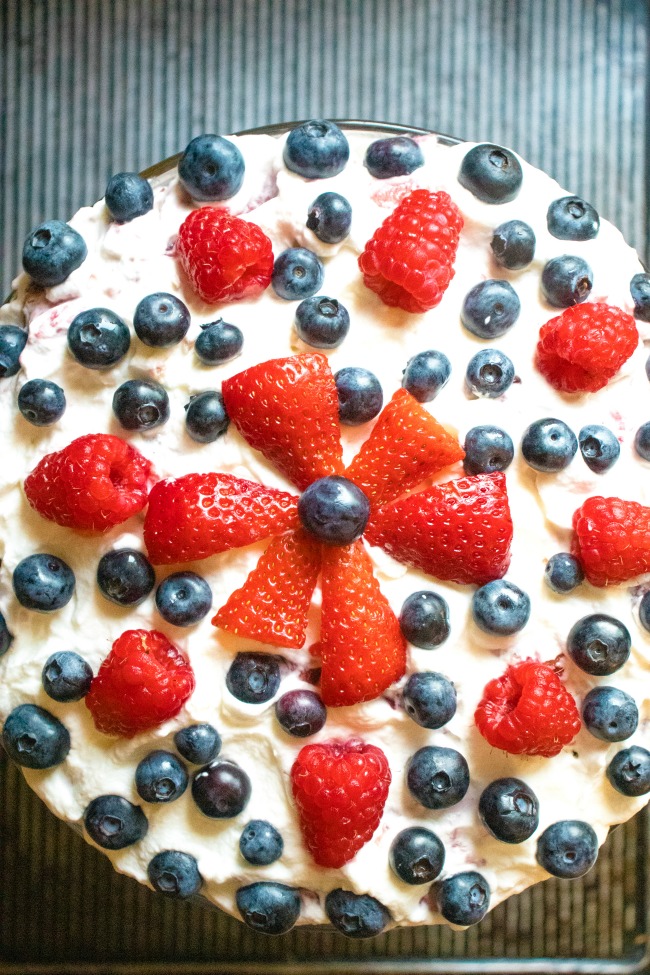 This photo features a above angle of a recipe called Raspberry Brandy Trifle. It is decorated with Strawberries, Raspberries, and Blueberries.