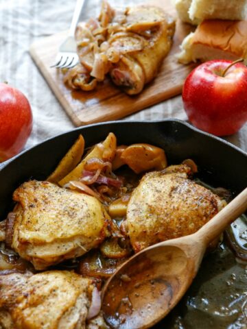 This photo features apple cider chicken in a skillet. Off to the side is a couple of apples and one of the thighs placed on a tray.
