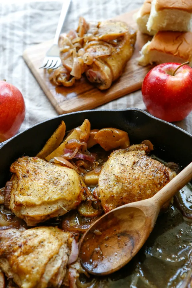 This photo features apple cider chicken in a skillet. Off to the side is a couple of apples and one of the thighs placed on a tray.