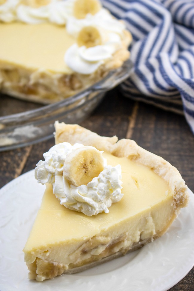 This photo features a slice of Banana Cream Pie on a white plate. It is topped with a dollop of whipped cream and slice of banana.