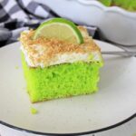 This photo features a piece of Key Lime Cake on a white plate. It has a slice of key lime as a garnish with some crushed graham crackers on top.