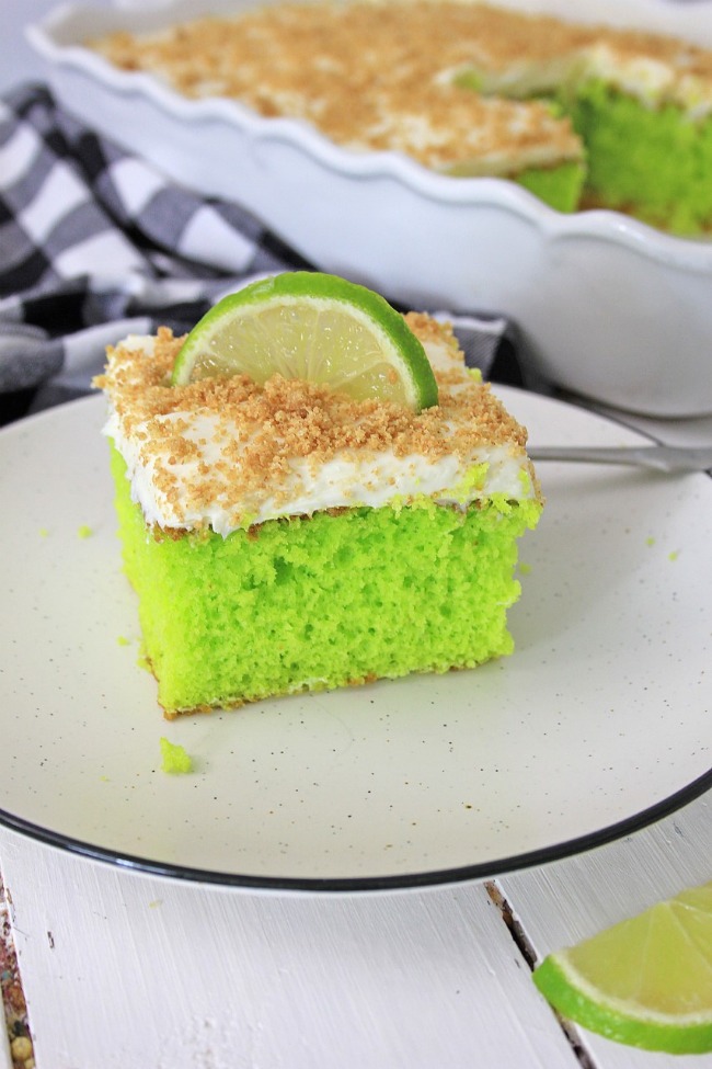 This photo features a piece of Key Lime Cake on a white plate. It has a slice of key lime as a garnish with some crushed graham crackers on top.