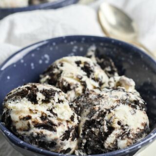 This photo features a blue bowl on a white tea towel. In the bowl is No Churn Oreo Ice Cream.