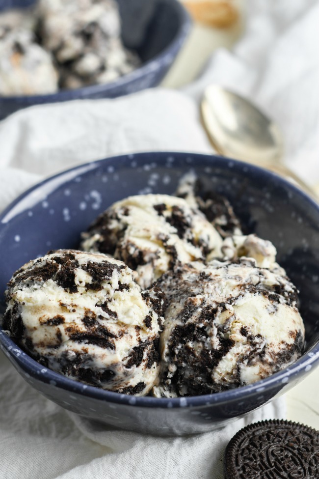 This photo features a blue bowl on a white tea towel. In the bowl is No Churn Oreo Ice Cream.