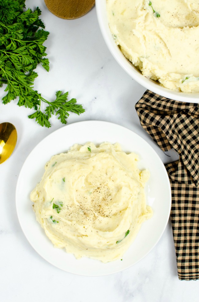This photo features a white plate of creamy and fluffy slow cooker mashed potatoes. Also in the picture are some photo props including parsley, a checkered tea towel and gold spoon.