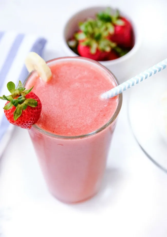 This photo features a glass of strawberry banana smoothie with a paper straw and a strawberry and banana on the rim. There is also a few side props, a tea towel and a dish of strawberries.