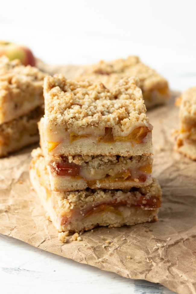 This photo features a stack of peach crumble bars sitting on a crumbled brown paper bag.