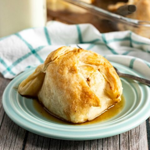 This photo features a side dish of apple dumplings with one single dumpling placed upon a blue saucer. In the background there is a white and blue checkered tea towel and a glass of milk.