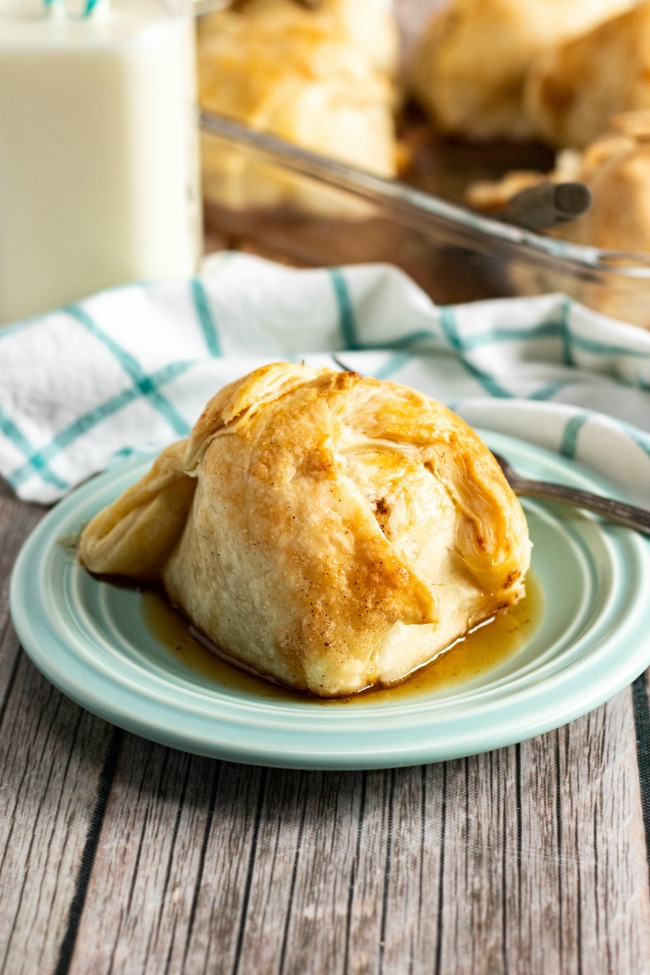 This photo features a side dish of apple dumplings with one single dumpling placed upon a blue saucer. In the background there is a white and blue checkered tea towel and a glass of milk.