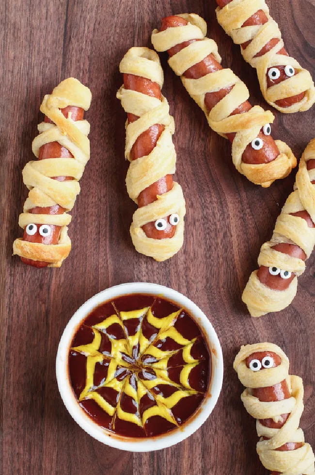 This photo features wrapped hot dogs in puff pastry. They are made to look like mummy dogs. On the side is a dish of ketchup and mustard. The mustard is swirled to look like a spider web.