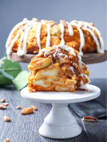 This photo features a Pumpkin Pecan Monkey Bread sitting in the background on a wooden pedestal. A slice of the bread has been taken out and on a smaller white pedestal in the foreground.