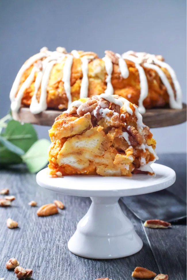 This photo features a Pumpkin Pecan Monkey Bread sitting in the background on a wooden pedestal. A slice of the bread has been taken out and on a smaller white pedestal in the foreground.