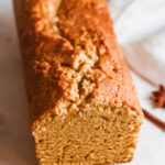 This photo features a loaf of pumpkin bread sitting on a white board with a white tea towel placed beside it and a cinnamon stick for prop.