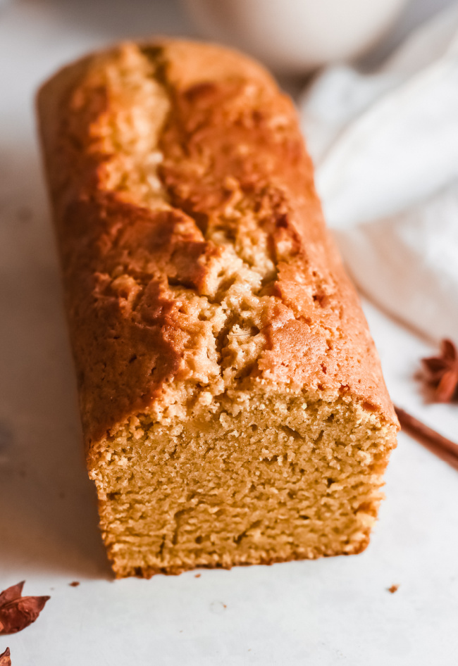 This photo features a loaf of pumpkin bread sitting on a white board with a white tea towel placed beside it and a cinnamon stick for prop.