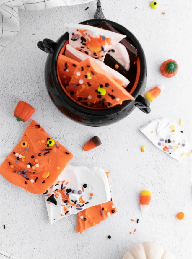 This photo features various versions of Halloween Candy Bark, some sitting on the table and others stacked in a cauldron candy dish.
