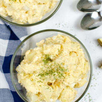 This photo features a bowl of the best potato salad around. In the background there is a blue and white tea towel and some spoons.