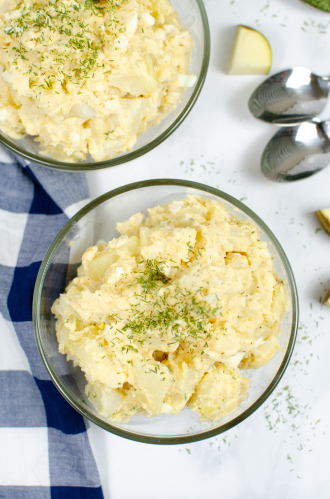 This photo features a bowl of the best potato salad around. In the background there is a blue and white tea towel and some spoons.