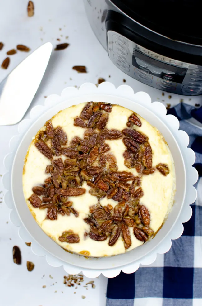 This photo features a Pecan Pie Cheesecake layed out on a white cake stand. A blue and white checkered tea towel is placed beside as a prop and in the background is an instant pot.