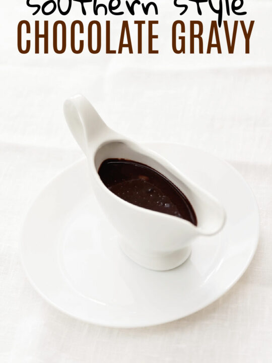 A white gravy boat filled with chocolate gravy sitting on a white plate with a white background