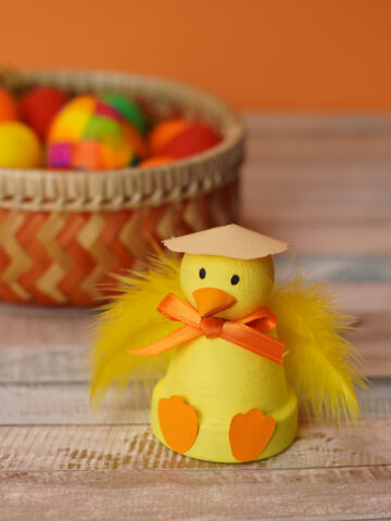 Easter chick terra cotta pot craft done up in yellow with a easter basket full of colorful eggs in back.