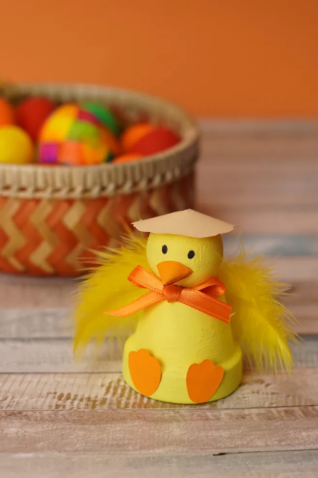 Easter chick terra cotta pot craft done up in yellow with a easter basket full of colorful eggs in back.