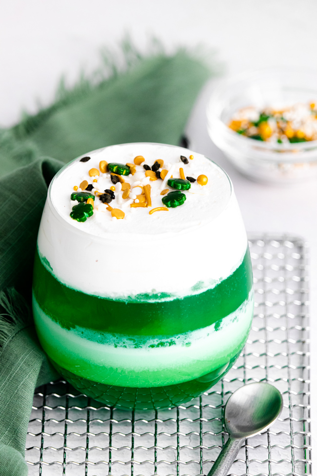 This photo features a glass cup of green jello mixed in with whipped topping to create St. Patrick's Day Jello Cups.