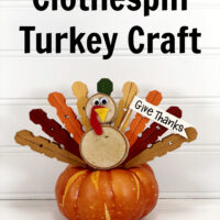 Clothespin Turkey craft sitting on top of a faux orange pumpkin with a white wood slate background.