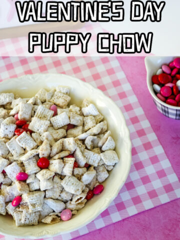 A white bowl full of Valentine's Day Puppy Chow on a pink and white gingham background