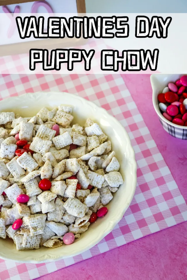 A white bowl full of Valentine's Day Puppy Chow on a pink and white gingham background