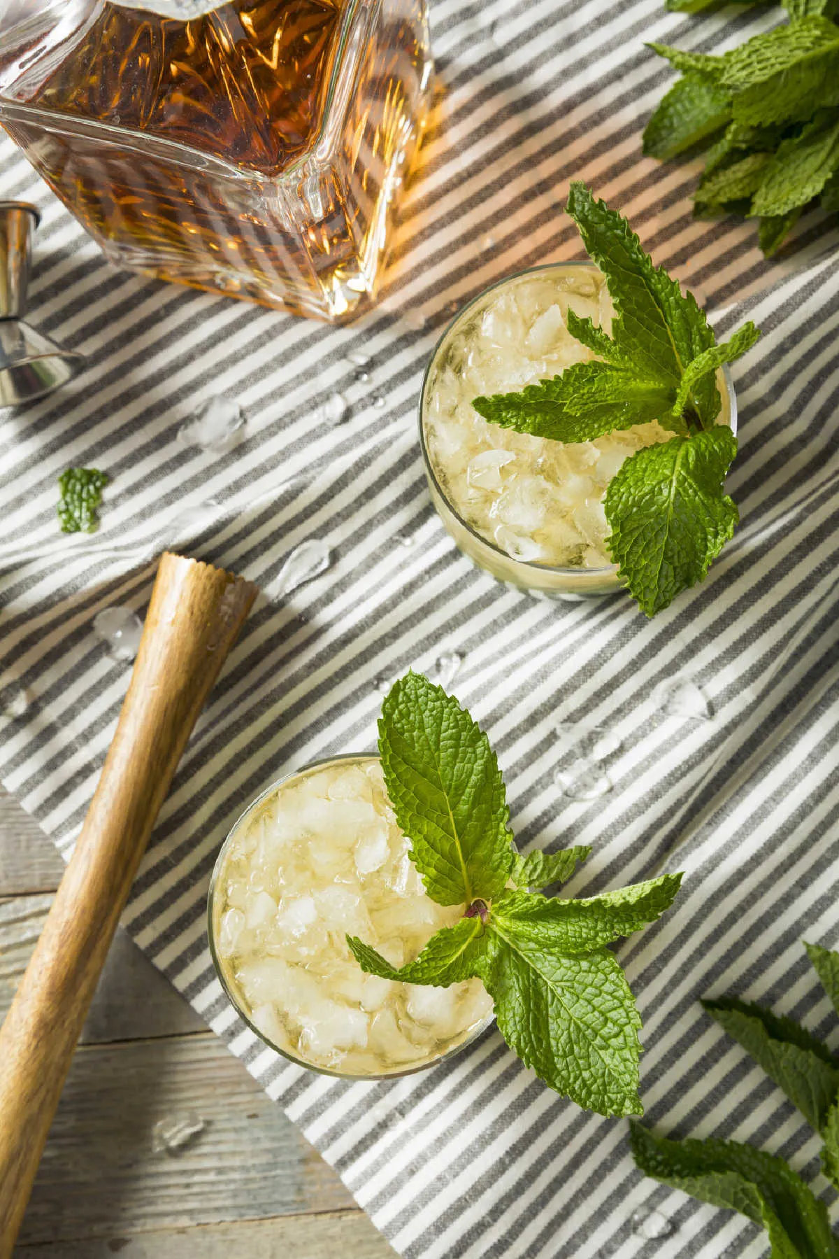 2 glasses of mint julep recipe with a canter of bourbon to the side and a muddler all laid out on a striped tea towel.