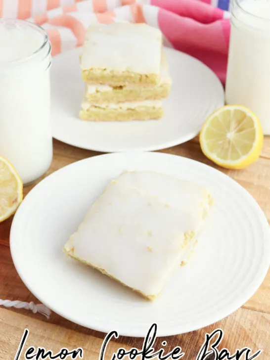 a white plate of lemon cookie bars with stacked cookie bars in the back and a sliced lemon on the side
