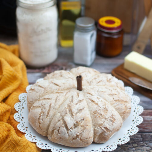 A pumpkin shaped bread on a white doily plate with a cinnamon stick placed in the top.